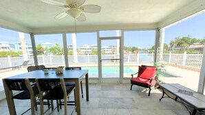 Área externa coberta com mesa e cadeiras, ventilador de teto e vista para uma piscina e edifícios.