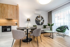 En moderne stue med et rundt spisebord, grå stole, en sofa, en grøn lænestol og et stort vindue med gardiner.