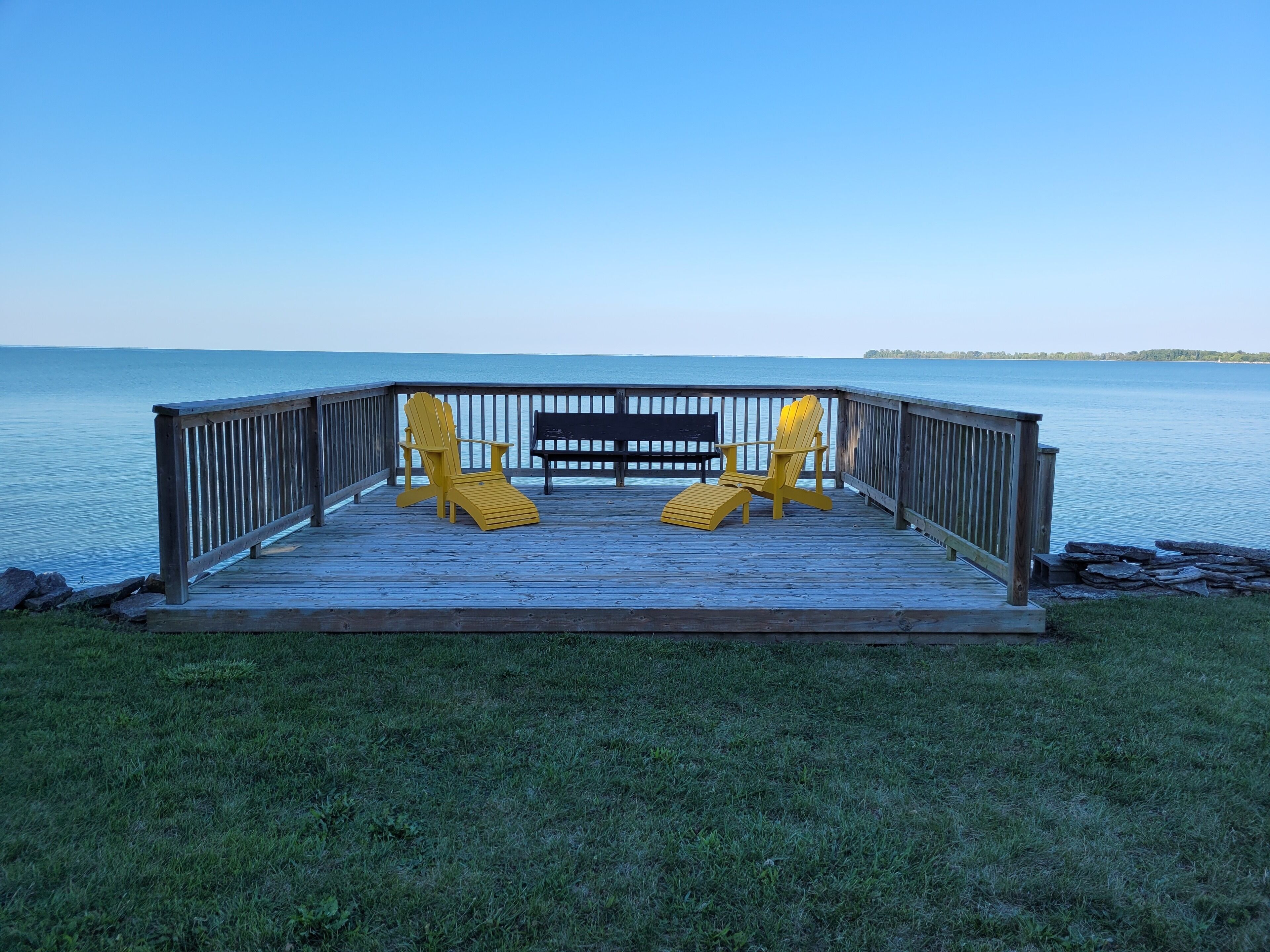 A wooden deck with two yellow Adirondack chairs and a black bench overlooking a calm body of water.