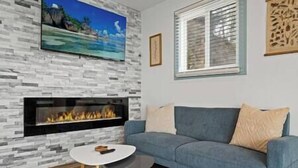 A living room with a stone-patterned wall, a fireplace, a blue sofa, a small round table, and a TV mounted on the wall. - Long Point Lake House - Daphne's House (Aurora)