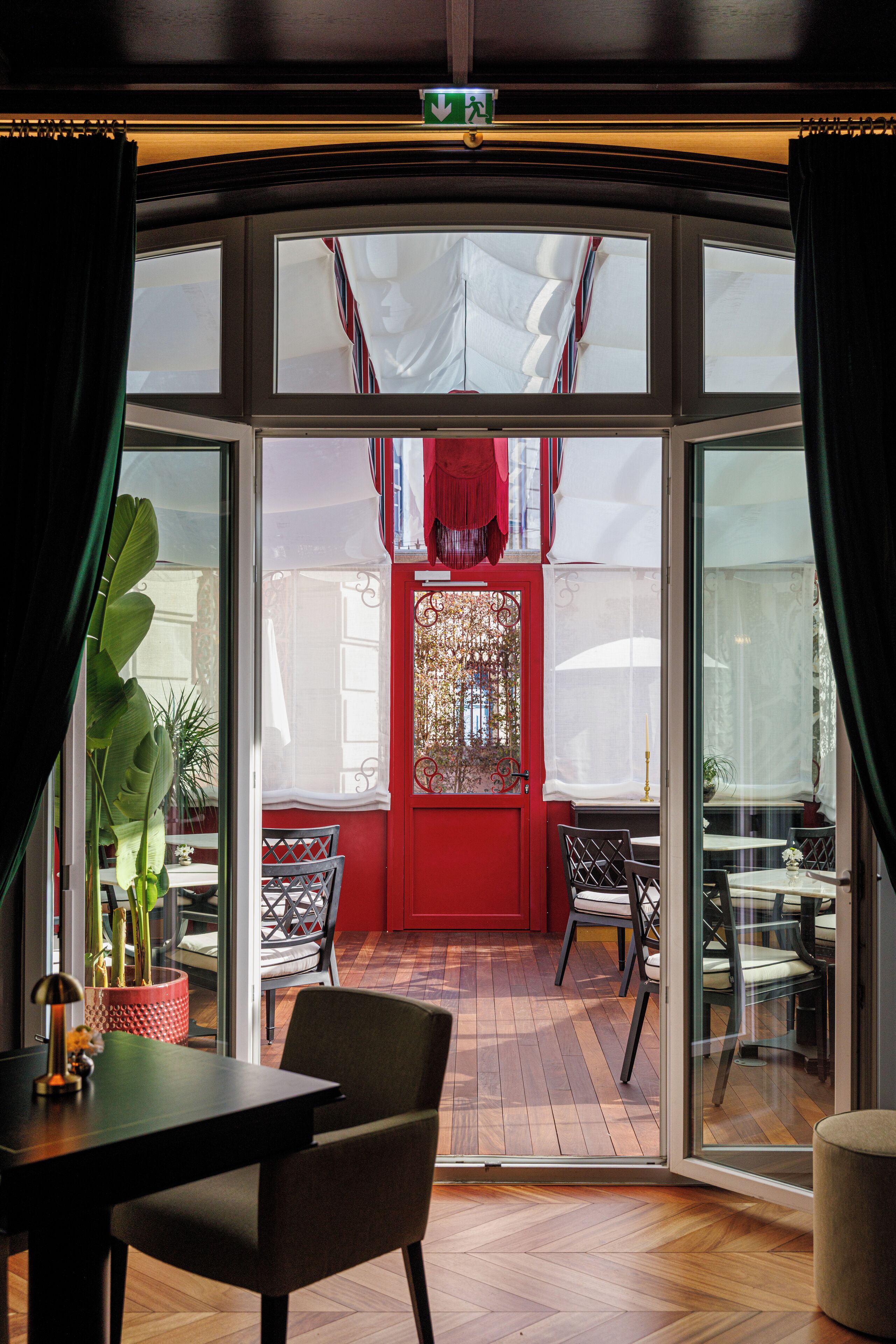 Una habitación con suelo de madera, una mesa, sillas, una planta en maceta y una puerta roja con paneles de vidrio decorativos.
