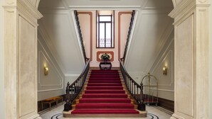 A grand staircase with a red carpet, flanked by white columns and a decorative ceiling.