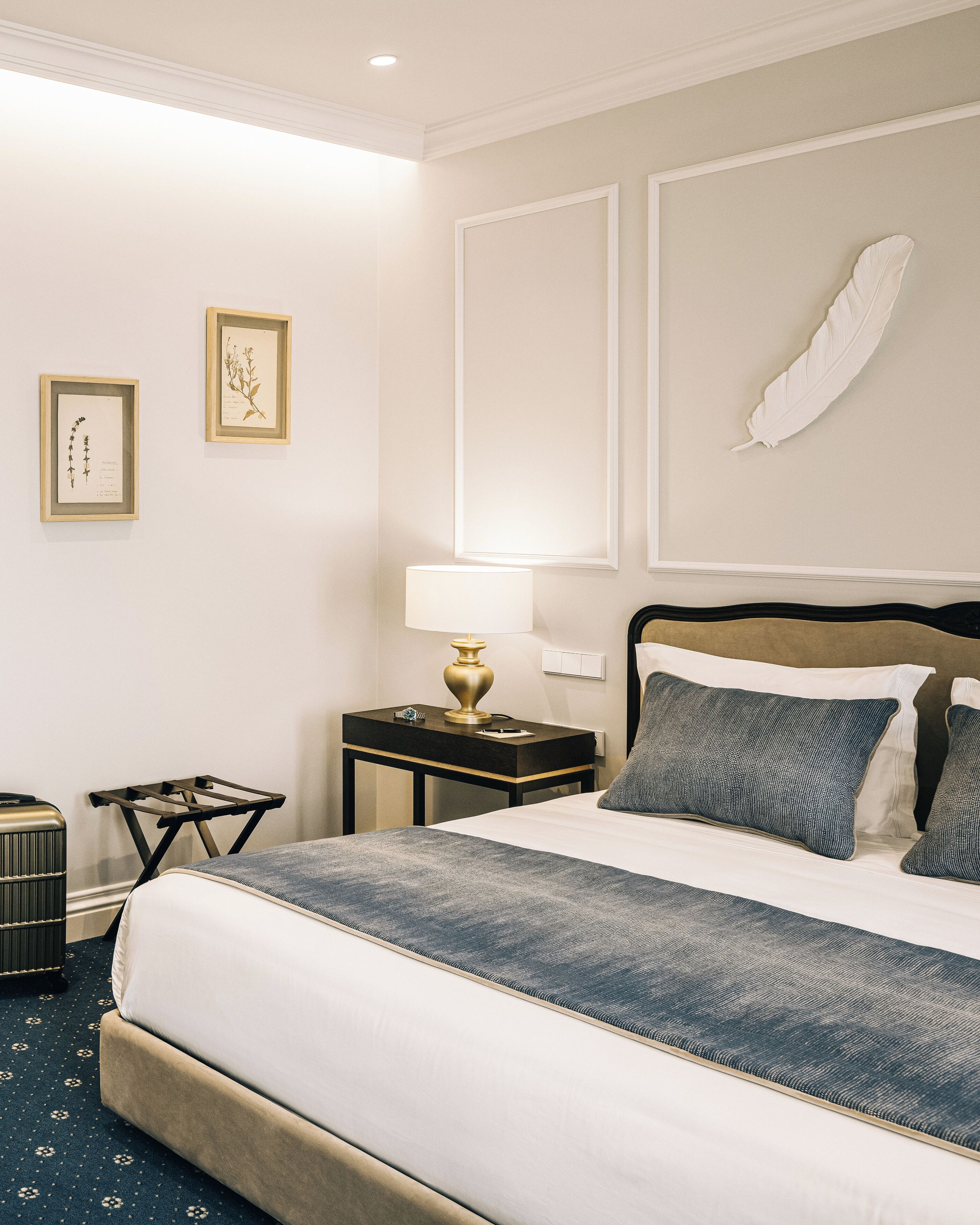 Habitación de hotel con una cama, una mesita de noche con lámpara, una maleta e ilustraciones botánicas enmarcadas en la pared.