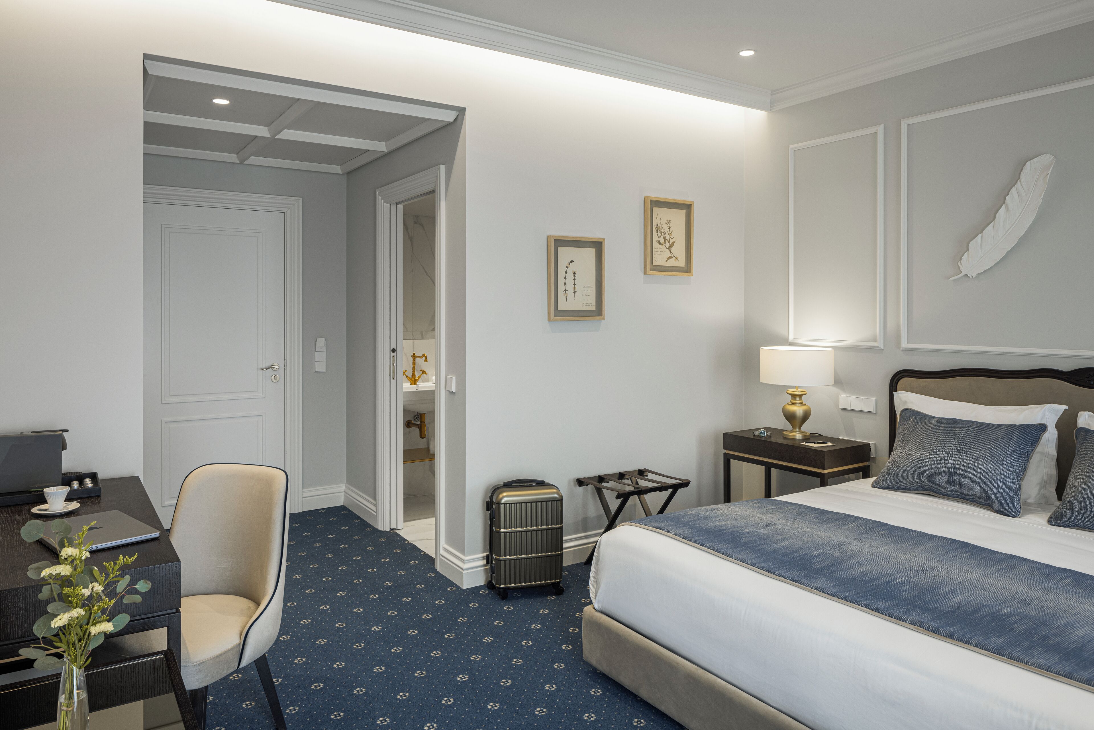Habitación de hotel con cama, escritorio con una computadora portátil, silla, maleta y cuadros en la pared.