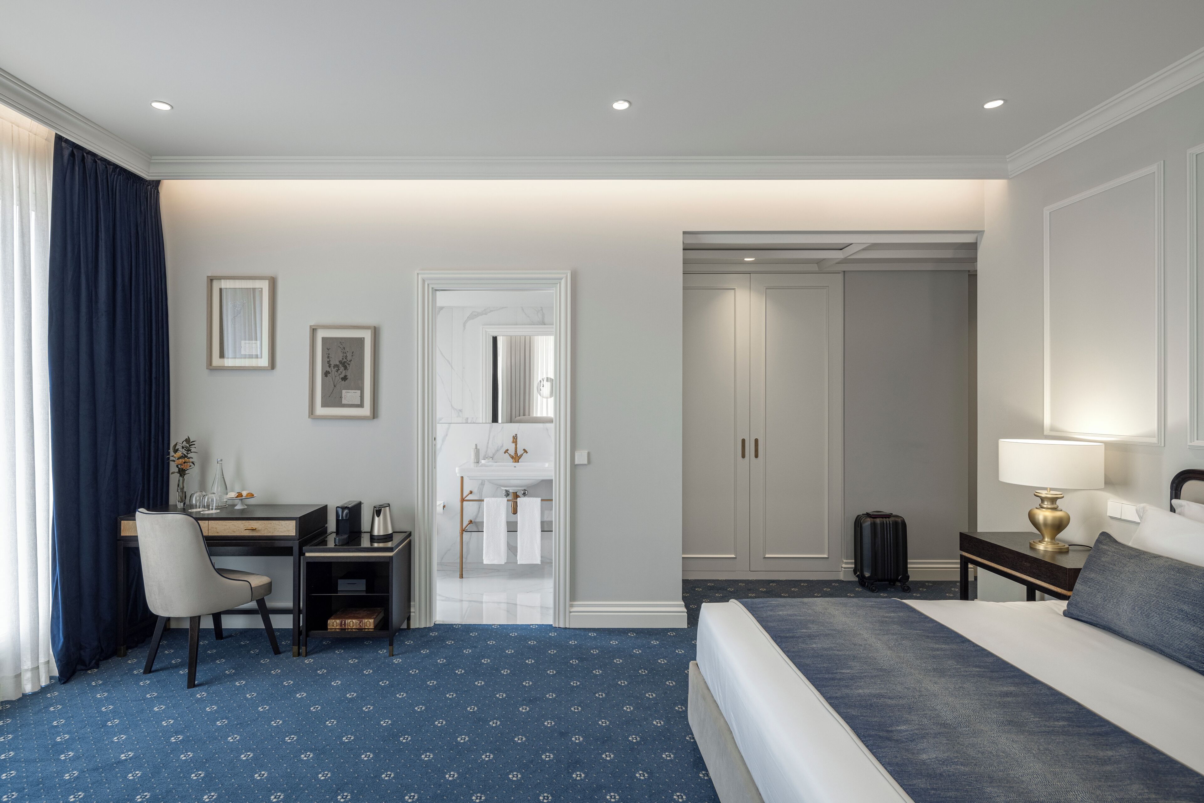 Habitación de hotel con cama, escritorio, silla, lámpara de escritorio, maleta y baño visible a través de una puerta abierta.