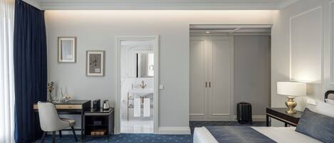 Habitación de hotel con cama, escritorio, silla, lámpara de escritorio, maleta y baño visible a través de una puerta abierta.