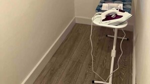 A corner room with a wooden floor, a window with blinds, and an ironing board with an iron. - Spacious Luxury 2 Bedroom Apartment, Station 1min (London)