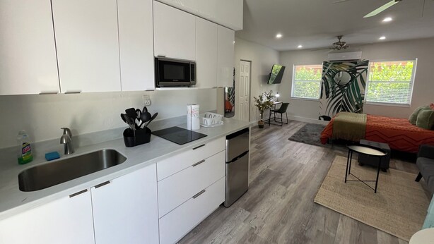 A modern kitchen with white cabinets, a stainless steel sink, and a microwave. There is a dining area with a table and chairs, and a bed with an orange bedspread. - Studios Casa Habana (Miami)