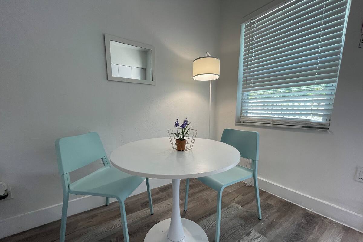 A dining area with a round table, two chairs, a wall-mounted mirror, a floor lamp, and a window with blinds.