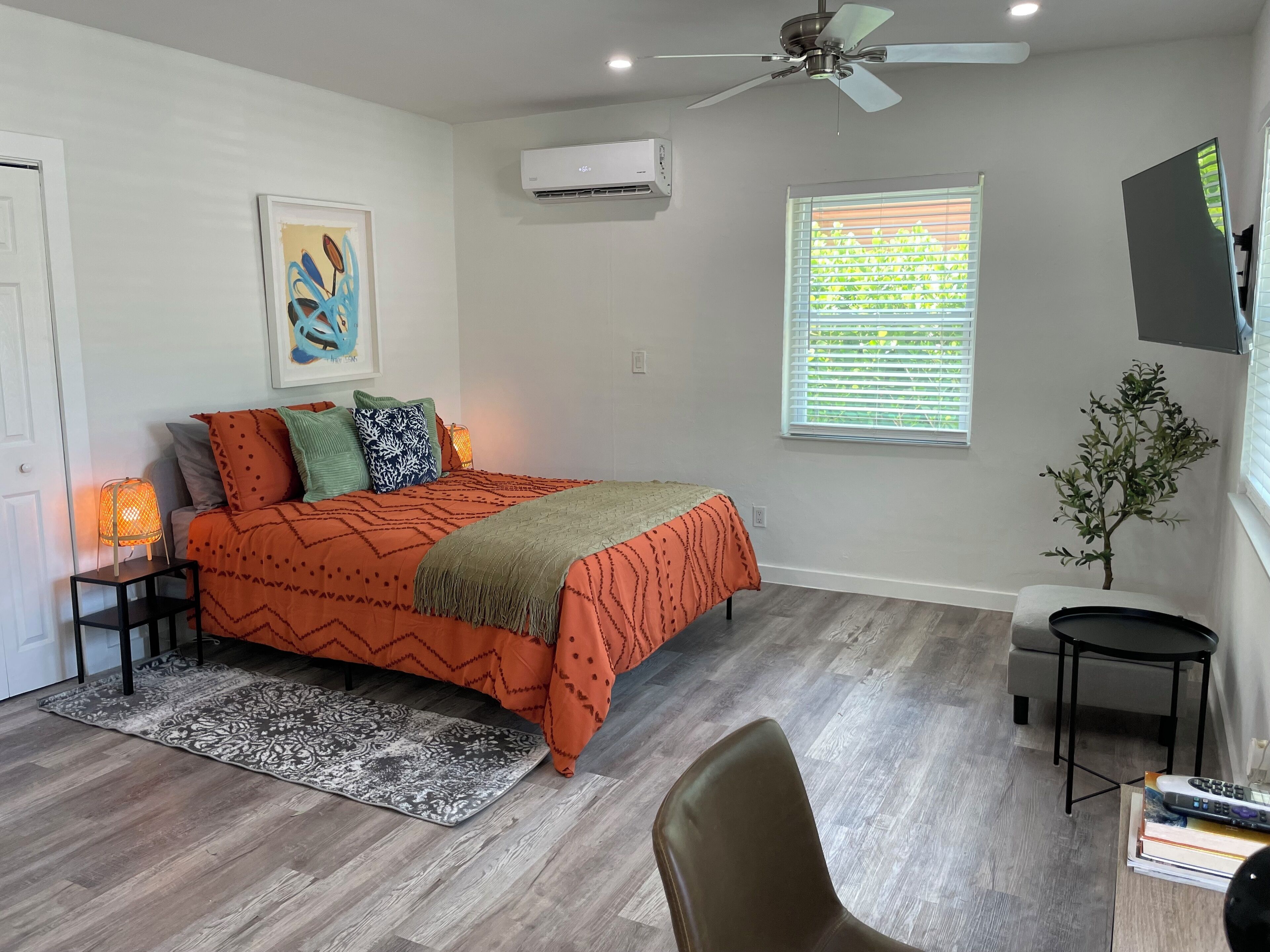 A bedroom with a bed, a nightstand, a chair, a small table, a window with blinds, an air conditioner, and a ceiling fan.
