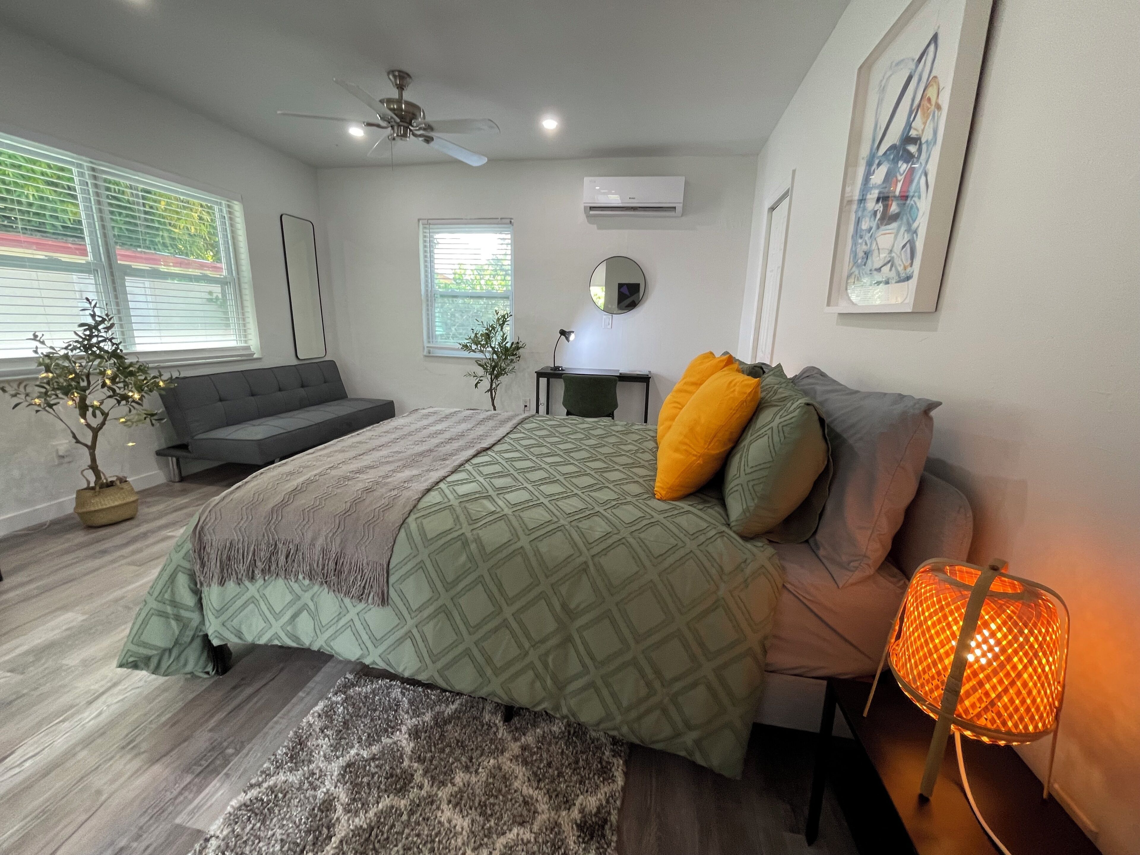 A modern bedroom with a bed, a ceiling fan, an air conditioner, a small table, and a window with blinds.