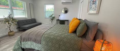 A modern bedroom with a bed, a ceiling fan, an air conditioner, a small table, and a window with blinds.