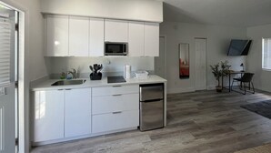 A modern kitchen with white cabinets, a stainless steel refrigerator, and a microwave. There is a dining area with a table and chairs, and a plant on the windowsill. - Studios Casa Habana (Miami)