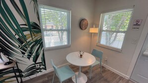 A room with a round white table, two light blue chairs, and a potted plant on the table. There are two windows with blinds, a wall-mounted lamp, and a 'No Smoking' sign. - Studios Casa Habana (Miami)
