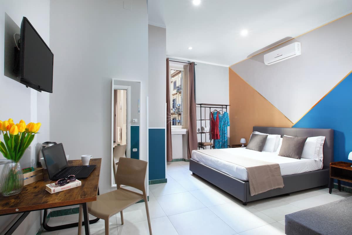 A bedroom with a bed, bedside table, and a desk with a laptop and a vase of flowers.
