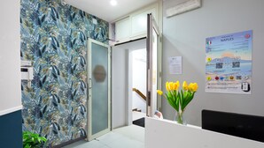 A reception area with a patterned wall, a glass door, a poster, and a vase with yellow flowers. - Royal Rooms Station Napoli (Naples)