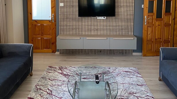 Ein Wohnzimmer mit Flachbildfernseher an einer Holzwand, einem Glastisch, zwei Sofas, einer Deckenleuchte und einer Tür mit Fenster.