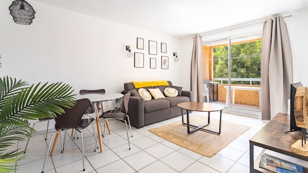 A modern living room with a grey sofa, a dining table with chairs, a coffee table, and a large window with curtains. - La Castillane - Appt avec piscine partagée (Cavalaire-sur-Mer)