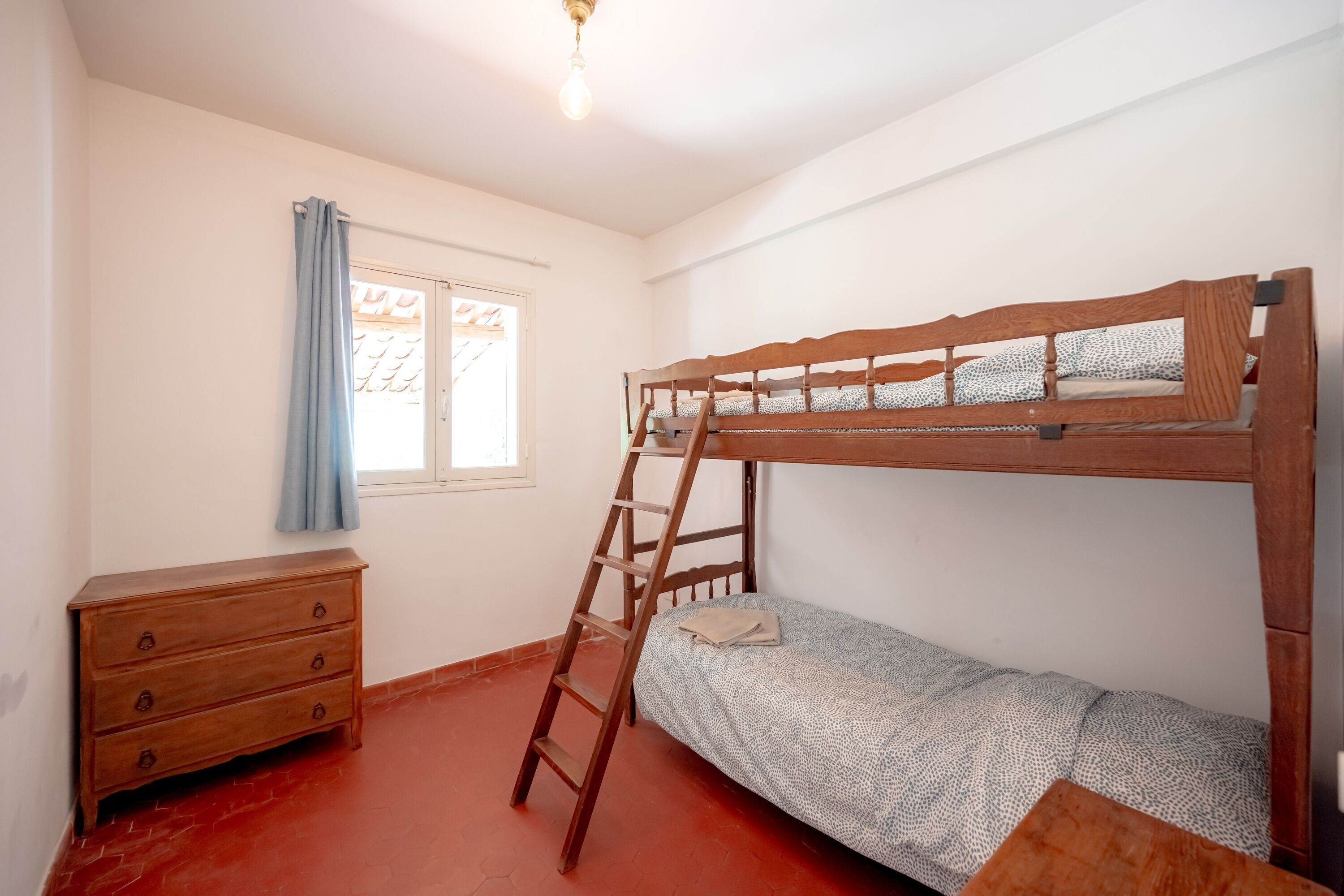 A single-bedded room with a wooden bunk bed, a wooden dresser, a ladder, a window with a curtain, and a light fixture.