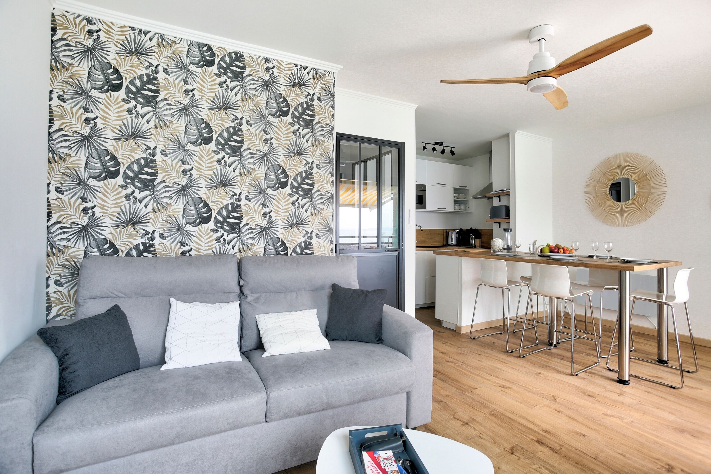 A modern living room with a grey sofa, a wooden dining table with white chairs, a ceiling fan, and a kitchen in the background.