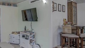 Quarto com televisão presa na parede, um armário pequeno com diversos objetos, uma mesa de madeira com cadeiras e uma porta que dá para outro cômodo.