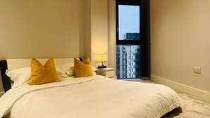 A bedroom with a large bed, two yellow pillows, a nightstand with a lamp, and a view of a building through a window.