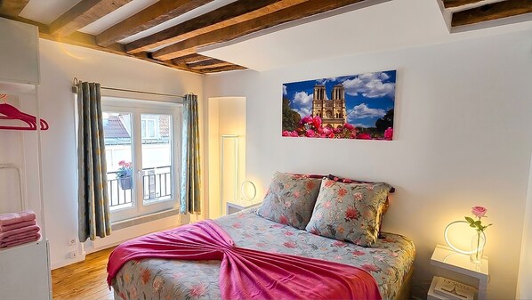 Un dormitorio con cama, ventana con cortinas, un cuadro de un edificio y una mesita de noche con un jarrón.