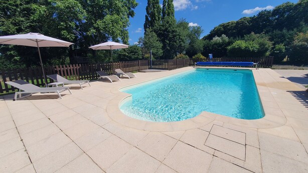 A rectangular swimming pool with a curved edge, surrounded by a tiled deck. There are lounge chairs and umbrellas along the poolside. - Studio "nid Cosy" Avec Piscine Chauffée (Bonnemain)
