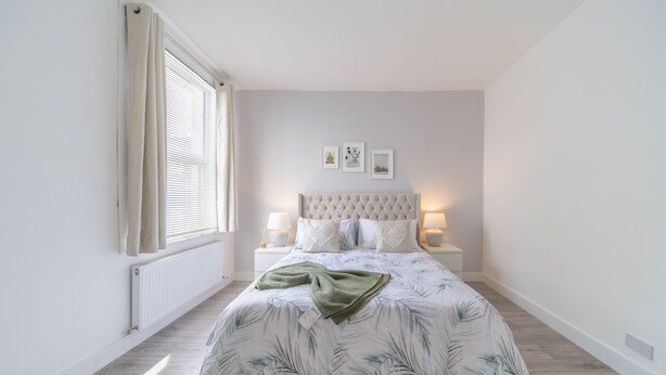 Une chambre à coucher avec une tête de lit capitonnée, un lit recouvert d’une couette à motifs de feuilles, deux lampes de chevet et un radiateur.