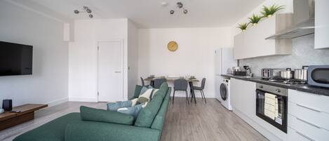 A modern living room with a green sofa, a flat-screen TV, and a dining area with a table and chairs.