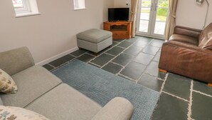 Uma sala de estar com sofá, apoio para os pés e um sofá de couro marrom. Há uma televisão sobre um suporte de madeira e uma janela com cortinas.