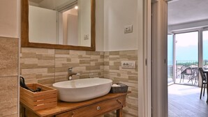 A bathroom with a wooden vanity, a large mirror, and a white sink. There is a wicker basket under the vanity and a door leading to a balcony with outdoor furniture. - Appartamento Lucia by Theater Home - with big balcony and a panoramic view (Tignale)