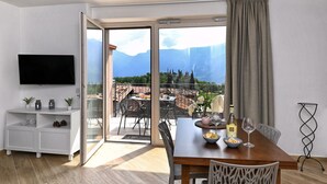 A modern room with a wooden dining table, chairs, a TV on the wall, and a balcony with a view of mountains. - Appartamento Lucia by Theater Home - with big balcony and a panoramic view (Tignale)