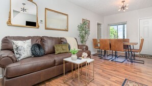 Ein Wohnzimmer mit einem braunen Ledersofa, zwei spiegeln mit goldfarbenen Rahmen, einem kleinen weißen Tisch mit einer Vase und einem Essbereich mit einem Holztisch und Stühlen.