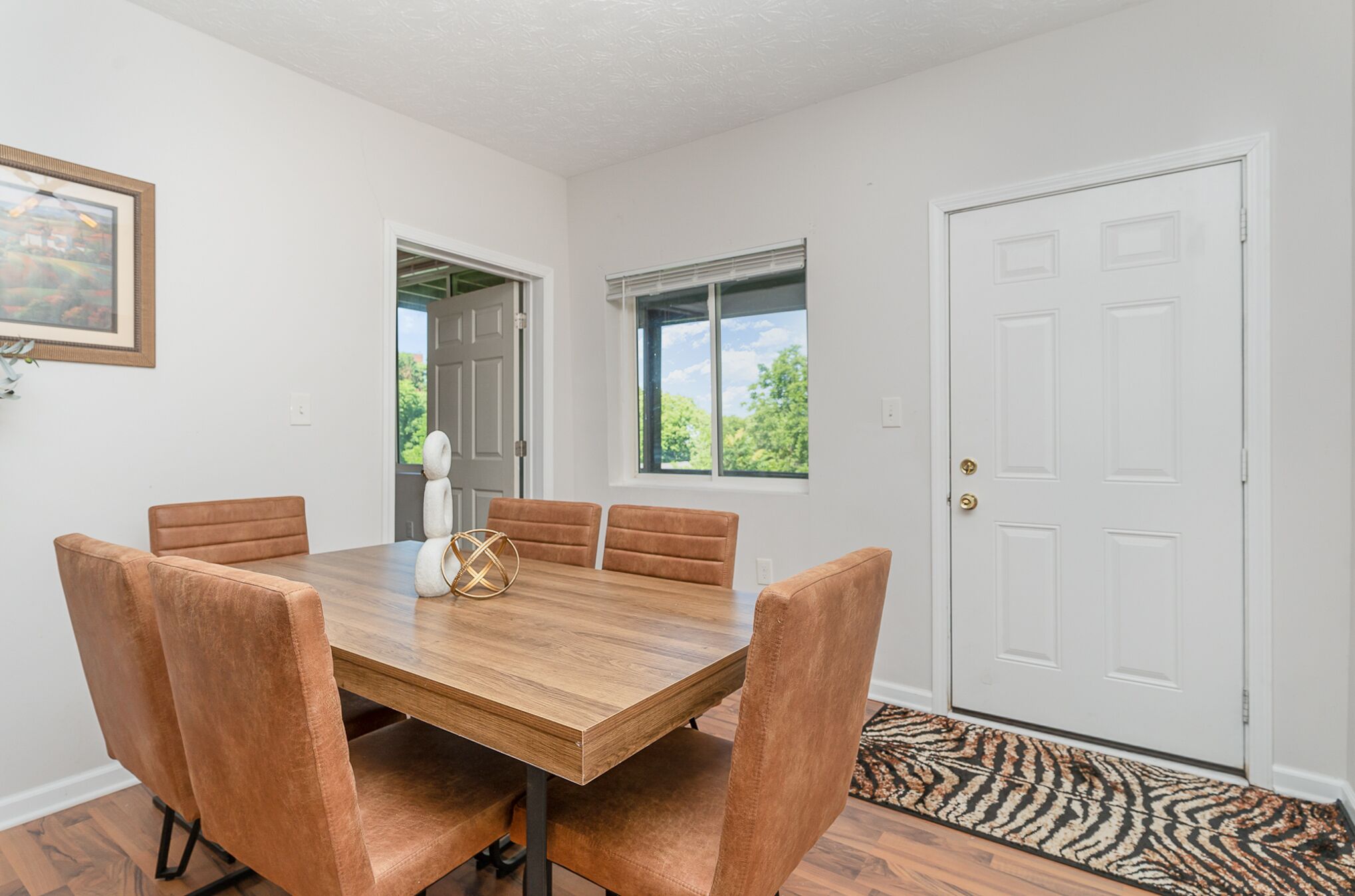 A dining room with a wooden table and six cushioned chairs, a framed picture on the wall, and a door with a doormat.