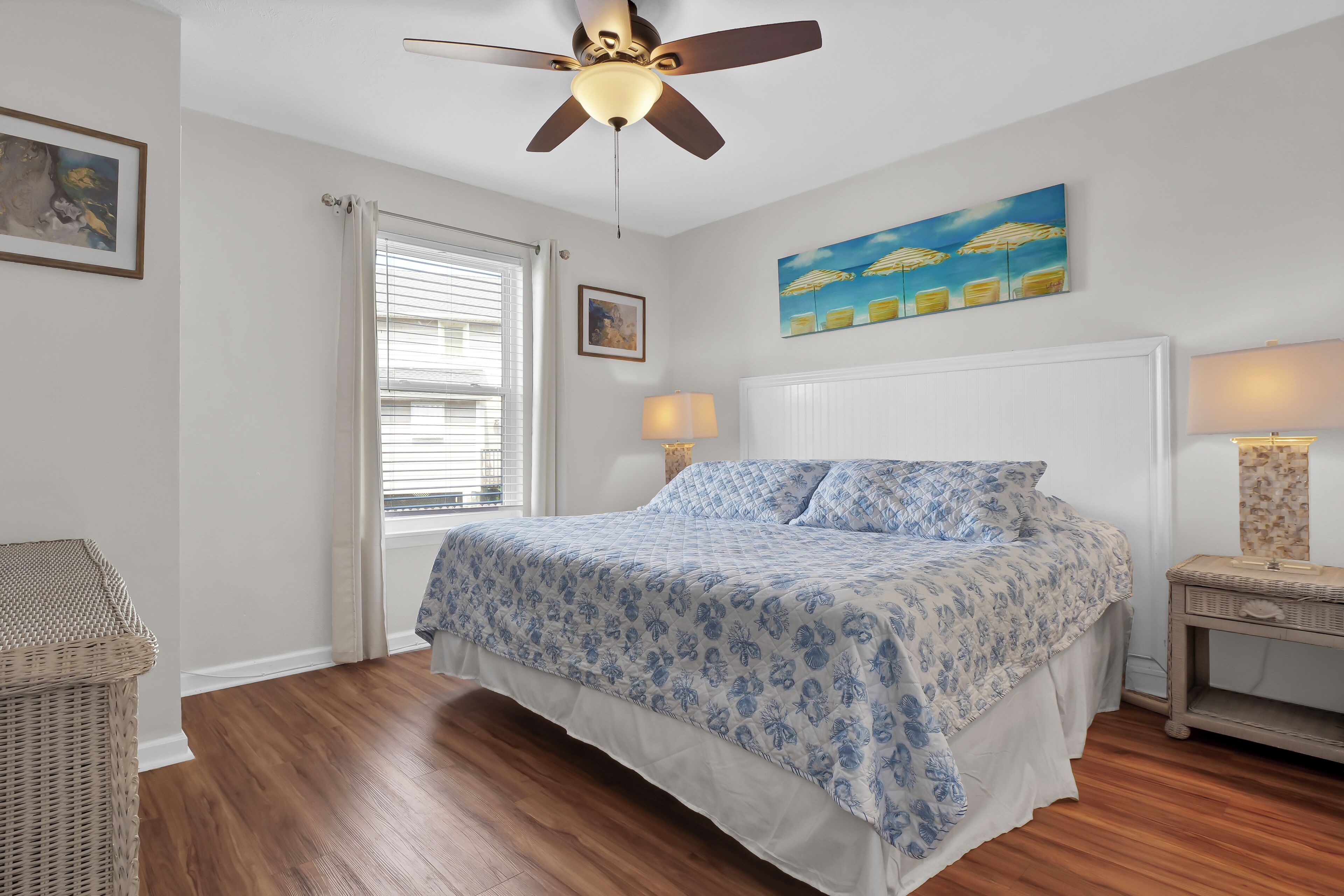 A bedroom with a bed, ceiling fan, window with blinds, wall art, and a bedside table with a lamp.