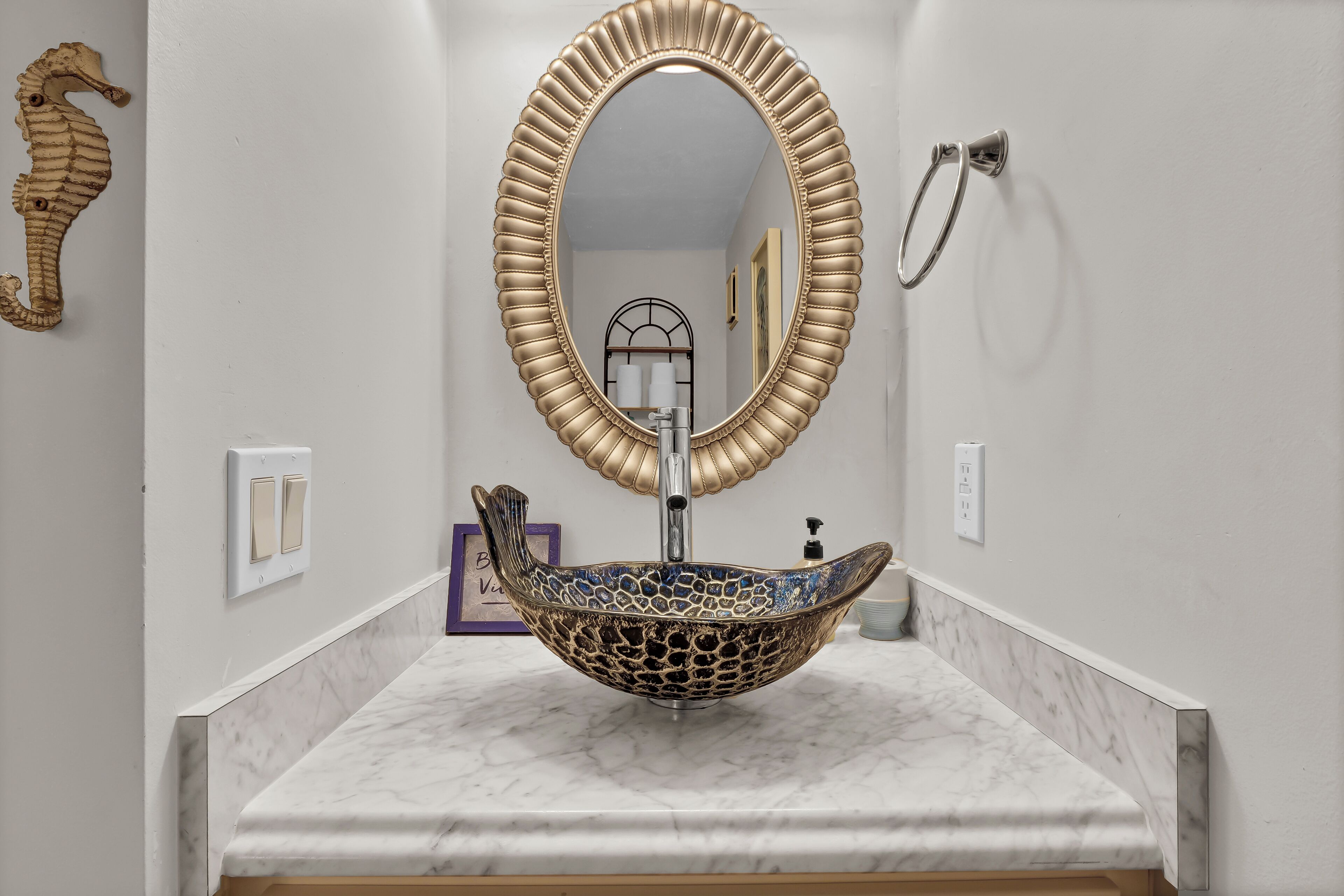 A bathroom with a marble countertop, a decorative seahorse, a gold-framed mirror, and a vessel sink.