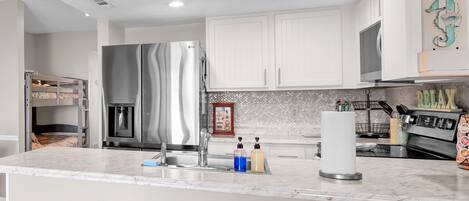 A modern kitchen with white cabinets, a stainless steel refrigerator, a sink, and a marble countertop. There are three stools with woven seats.
