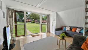 A spacious living room with a grey sofa, a small side table with a plant, and a large sliding glass door leading to a garden. - The Lookout, Sunny Beach Retreat, Sleeps 5 Guests (Deal)