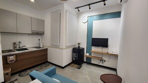 Une cuisine compacte équipée d’un évier, d’une cuisinière et de placards, un petit coin salon avec un canapé et une table, et un téléviseur fixé au mur.
