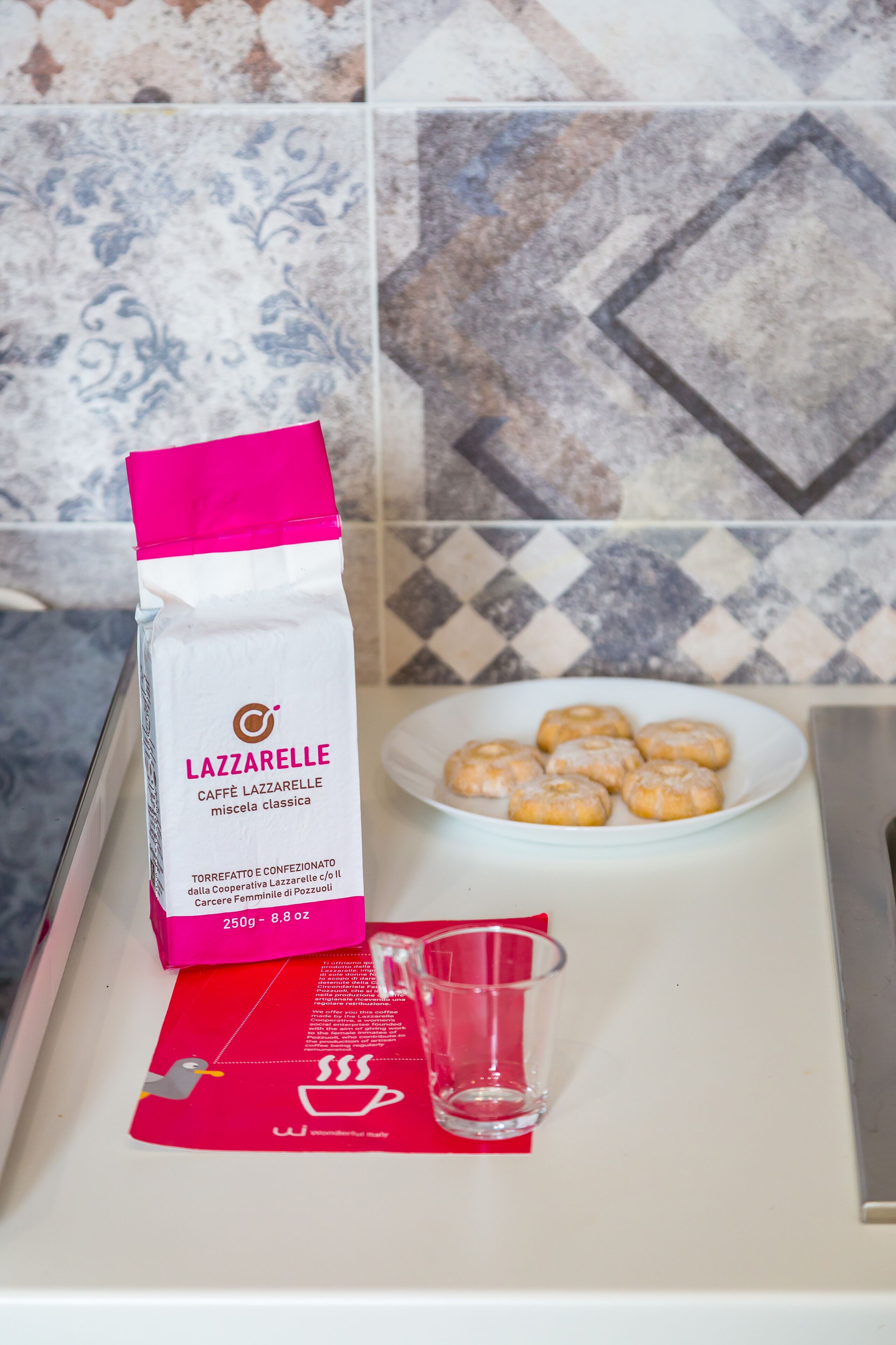 A kitchen counter with a bag of Lazzarelle coffee, a plate of pastries, and a glass.