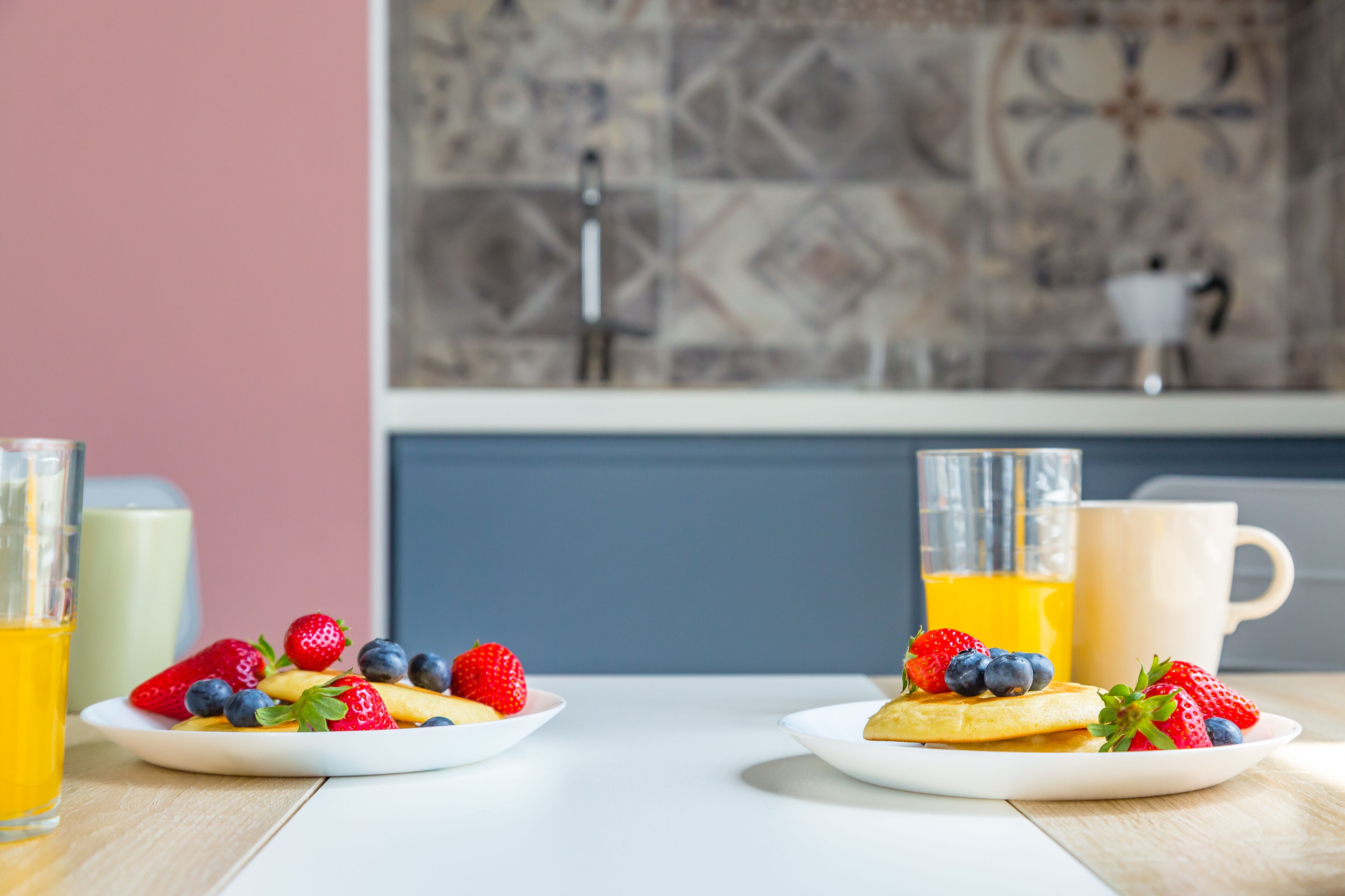 Two plates of pancakes with strawberries and blueberries, a glass of orange juice, and a mug on a table in a kitchen with a tiled backsplash and a sink.