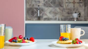 Two plates of pancakes with strawberries and blueberries, a glass of orange juice, and a mug on a table in a kitchen with a tiled backsplash and a sink.