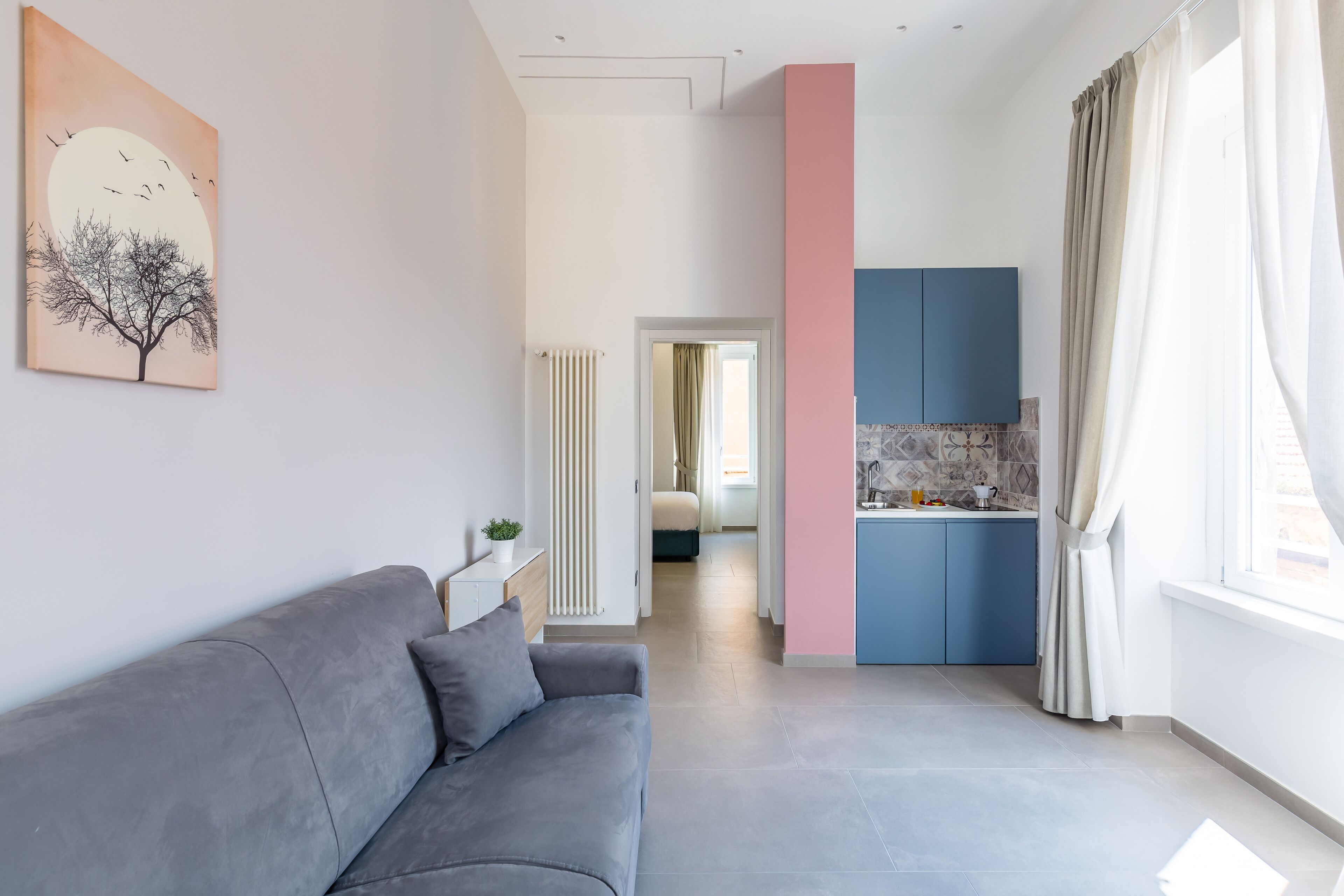 Een moderne woonkamer met een grijze bank, een schilderij van een boom en uitzicht op een keuken met blauwe keukenkastjes.