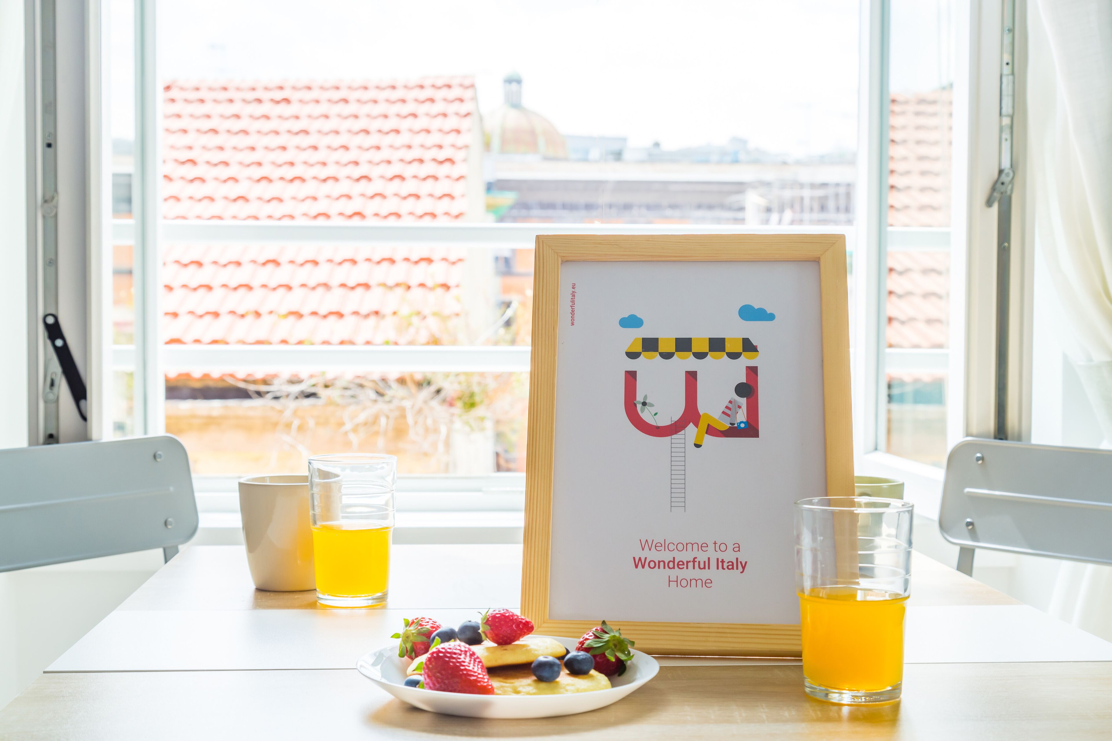Un panneau d’accueil encadré, orné d’une illustration de train, avec une assiette de fruits et un verre de jus d’orange posés sur une table près d’une fenêtre.