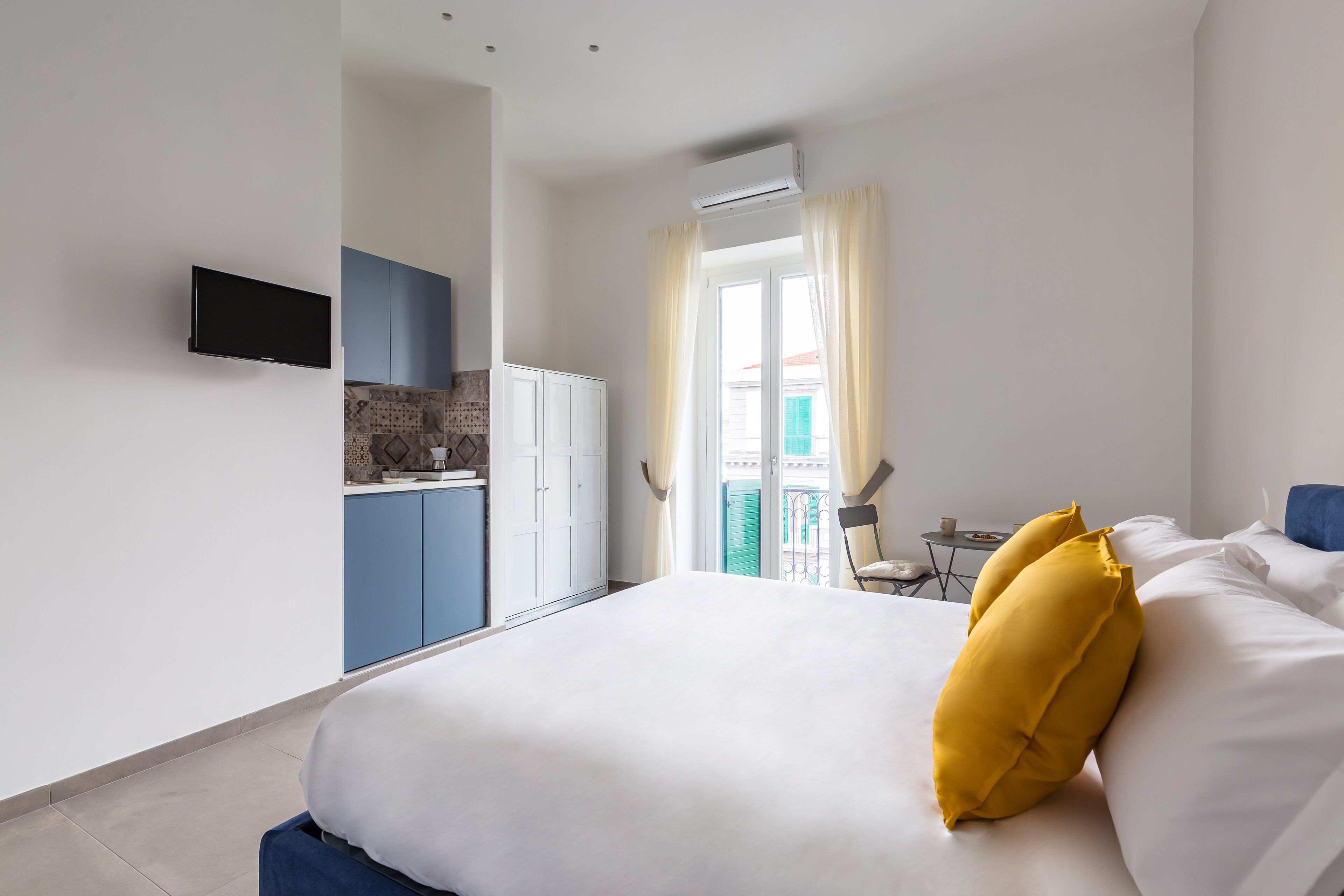 Habitación de hotel con cama, televisor montado en la pared, kitchenette y balcón con una silla.