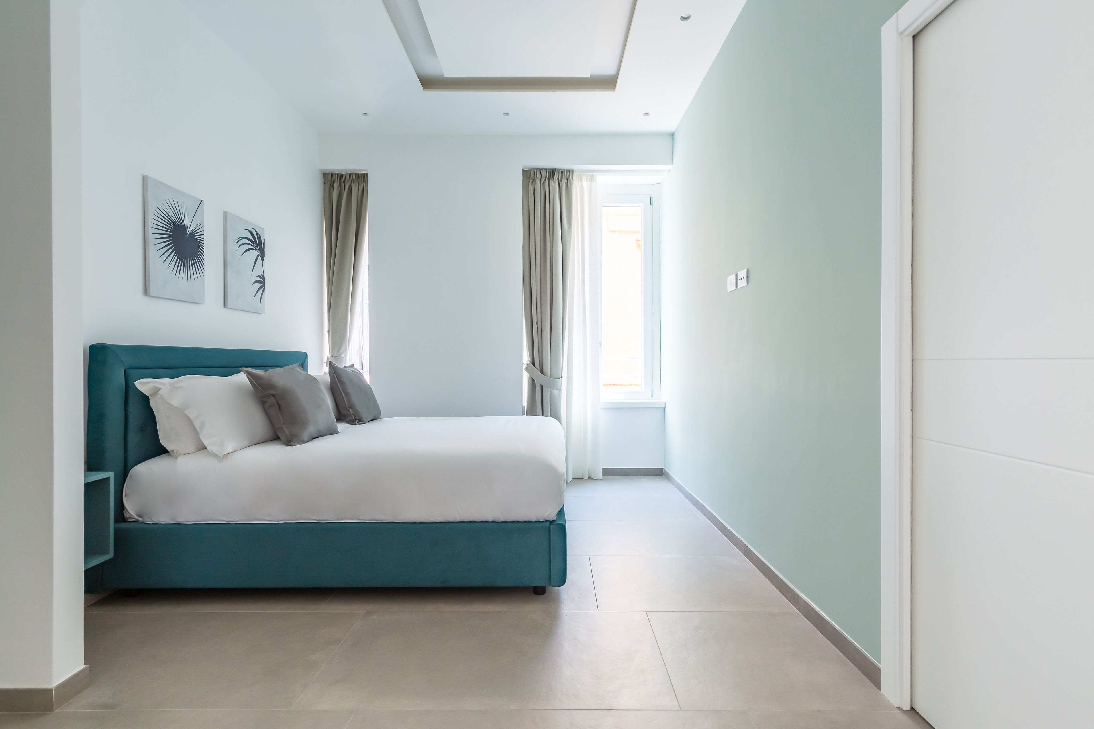 Een moderne slaapkamer met een blauwgroen bed, witte beddengoed en twee abstracte kunstwerken aan de muur.