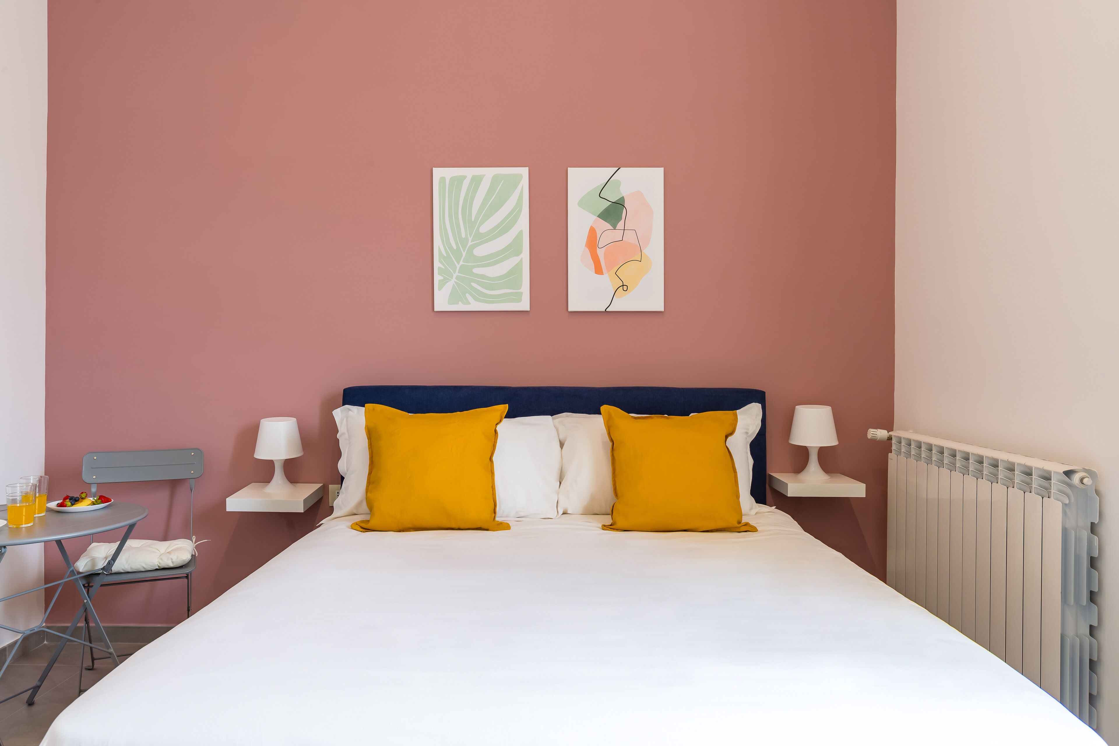 A neatly made bed with two yellow pillows, a bedside table with a lamp, and a chair with a plate of fruit.