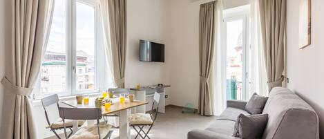 Una sala de estar moderna con un sofá, una mesa de comedor con sillas, un televisor de pantalla plana montado en la pared y ventanas amplias con cortinas.