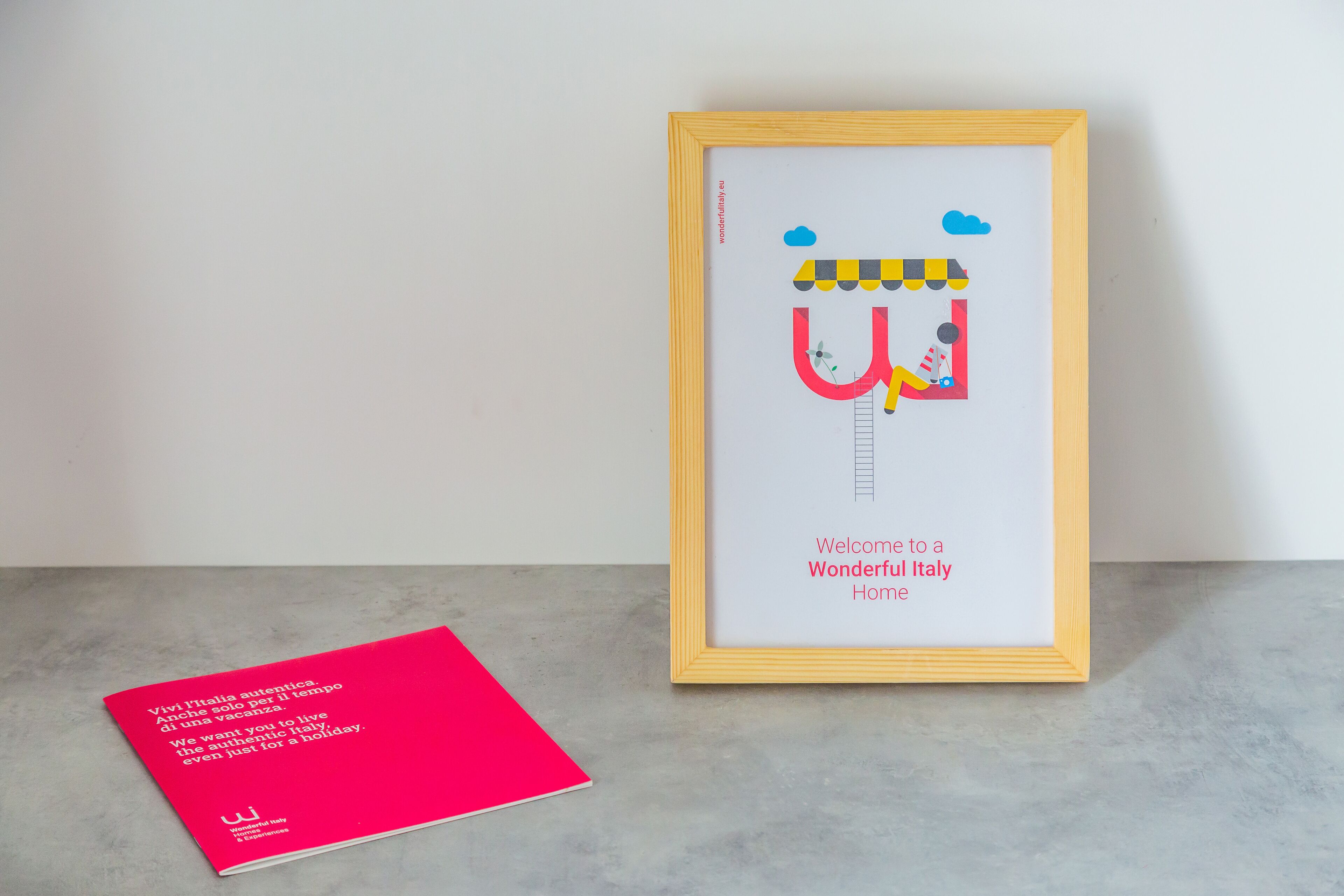 A framed picture with a welcome message to Italy, a red brochure, and a concrete floor.